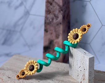 May include: A whimsical, turquoise-colored, spiral-shaped sculpture with three yellow sunflowers with brown centers. The sculpture rests on a beige rectangular block and a beige surface, with a pink crystal in the background.