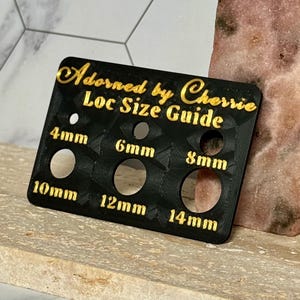 Puede incluir: Una gu&iacute;a de tama&ntilde;o de locs negra con letras doradas que dice "Adorned by Cherrie Loc Size Guide". La gu&iacute;a tiene agujeros etiquetados como 4 mm, 6 mm, 8 mm, 10 mm, 12 mm y 14 mm. La gu&iacute;a est&aacute; sobre una superficie clara.