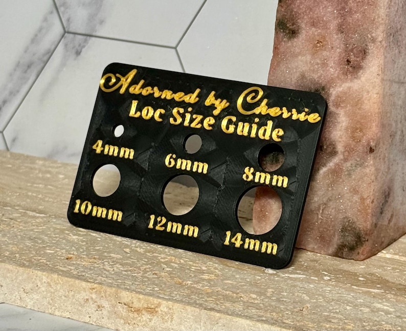 Puede incluir: Una gu&iacute;a de tama&ntilde;o de locs negra con letras doradas que dice "Adorned by Cherrie Loc Size Guide". La gu&iacute;a tiene agujeros etiquetados como 4 mm, 6 mm, 8 mm, 10 mm, 12 mm y 14 mm. La gu&iacute;a est&aacute; sobre una superficie clara.