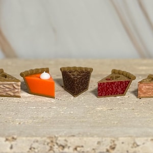May include: Five miniature pie slices with various fillings, including pumpkin, chocolate, and berry, are arranged on a light-colored surface. Each slice has a detailed crust and a distinct filling color, with one topped with a dollop of white cream.