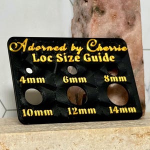 Puede incluir: Una gu&iacute;a de tallas de locs negra con letras doradas. La gu&iacute;a tiene seis agujeros de diferentes tama&ntilde;os, etiquetados 4 mm, 6 mm, 8 mm, 10 mm, 12 mm y 14 mm. El texto "Adorned by Cherrie Loc Size Guide" est&aacute; en la parte superior.