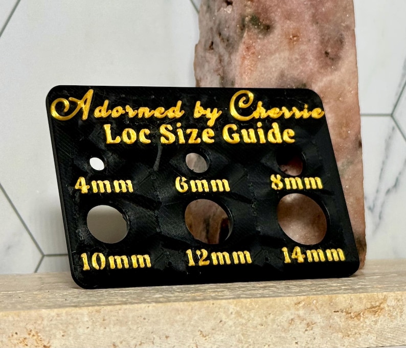 Puede incluir: Una gu&iacute;a de tallas de locs negra con letras doradas. La gu&iacute;a tiene seis agujeros de diferentes tama&ntilde;os, etiquetados 4 mm, 6 mm, 8 mm, 10 mm, 12 mm y 14 mm. El texto "Adorned by Cherrie Loc Size Guide" est&aacute; en la parte superior.