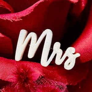 May include: A close-up of a red rose with the word "Mrs" in white script. The rose has a velvety texture, and the text is slightly raised, creating a 3D effect. The image is well-lit, highlighting the details.