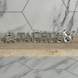 May include: A silver-colored metal sculpture of the word "TAURUS" with the Taurus zodiac symbol. The sculpture features a bull and the zodiac symbol. The letters are in a block style. The sculpture is displayed on a light brown surface.