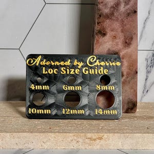 May include: A black Loc Size Guide with gold lettering that reads "Adorned by Cherrie". The guide has holes labeled 4mm, 6mm, 8mm, 10mm, 12mm, and 14mm. The guide is on a light brown surface.
