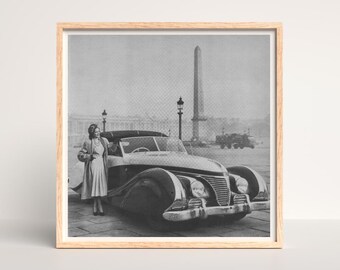 May include: Black and white photograph of a vintage convertible car with a woman standing beside it. The car has a sleek design with large fenders and a prominent front grille. The background features a tall obelisk and street lamps.
