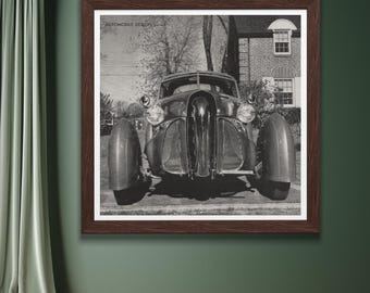 May include: Framed black and white print of a vintage car. The car has a long hood, rounded fenders, and a prominent grille. The print is in a dark brown frame and is mounted on a dark green wall.