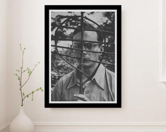 May include: Black and white framed photograph of a man wearing glasses, partially obscured by a metal structure. The man is wearing a button-down shirt. The photograph is displayed on a white wall.