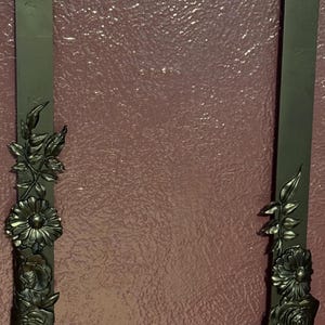May include: A dark green picture frame with a decorative floral design around the edge. The flowers are made of metal and are painted in a gold color.