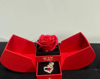 Può includere: Una scatola di velluto rosso con una rosa rossa e una collana con un ciondolo a forma di cuore all'interno. La scatola è aperta e la collana è visibile. Il testo sulla scatola dice "I migliori auguri Essere con te".