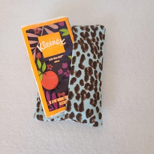 May include: A Kleenex On the Go ultra tissue pack is nestled in a light blue fabric holder with a brown leopard print. The tissue box has a floral design with an orange fruit.