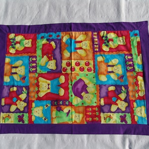May include: A colorful quilt with a patchwork design featuring teddy bears in various poses and colors. The quilt has a purple border and is made of soft fabric.