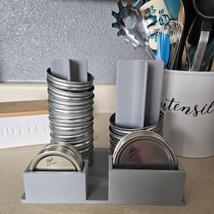 May include: A gray organizer holds stacks of silver jar lids and gray dividers. The organizer has two sections for lids and two sections for dividers. A white utensil holder with black text is in the background.