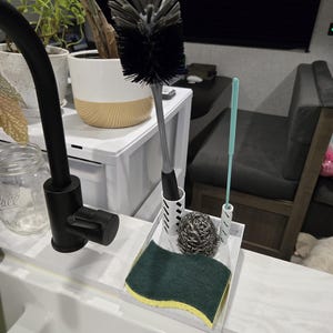 Puede incluir: Un área de fregadero de cocina con productos de limpieza. Un grifo negro está a la izquierda. Una bandeja blanca contiene un cepillo para botellas, una esponja verde y amarilla, un estropajo de lana de acero y una varilla de limpieza azul.