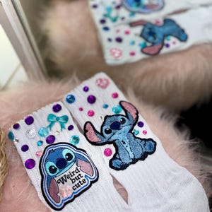 May include: White socks with blue and pink Stitch character patches and colorful gem embellishments. One sock features the text "Weird but cute". The socks are decorated with blue bows and pink and purple gems.