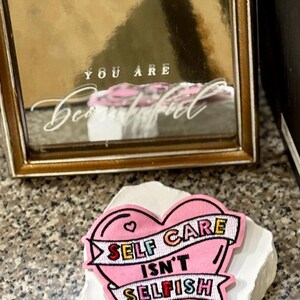 May include: A gold-framed mirror with the words "YOU ARE Beautiful" and a pink heart-shaped patch that reads "SELF CARE ISN'T SELFISH" on a white stone. The patch has a black outline and colorful text.