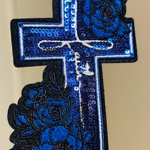 May include: A blue sequined cross patch with a black border and floral accents. The cross features the word "faith" in white script. The design includes blue roses and is suitable for apparel or craft projects.