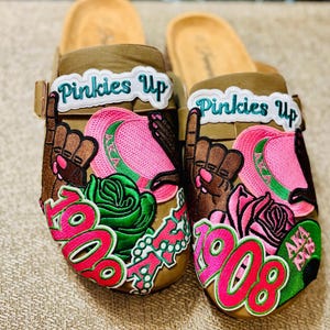 May include: Brown clogs with decorative patches. The patches include the text "Pinkies Up", a hand with the pinky finger raised, roses, and the numbers "1908" in pink and green. The clogs have a light brown footbed and a buckle strap.