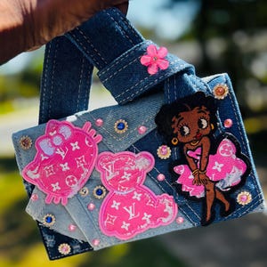 Könnte beinhalten: Jeans-Handtasche mit rosa und weißen Aufnähern von Schleife, Teddybär und Betty Boop. Die Tasche ist mit Strasssteinen und einer rosa Blume verziert. Der Griff ist aus Jeansstoff.