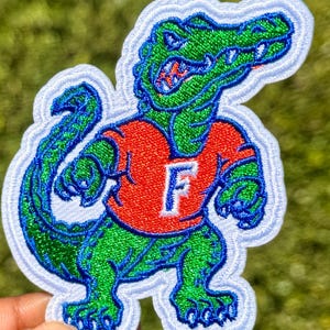May include: An embroidered patch featuring a green alligator character wearing a red shirt with a white letter F on the chest. The alligator has blue outlines and is set against a white background with a thick white border around the entire shape.