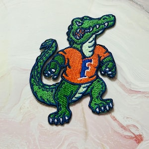 May include: Embroidered patch featuring a green alligator wearing an orange shirt with a blue "F" on the front. The alligator has white teeth and claws, with blue outlines. The patch is likely for apparel or accessories.