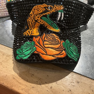 May include: A black visor covered in small black rhinestones. The front features an embroidered patch of a snake with orange and green details, positioned above a large orange rose flanked by two smaller green roses on either side of the brim.