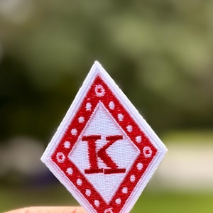 May include: A diamond-shaped embroidered patch featuring a white background with a red border. The border is decorated with a repeating white dot pattern. Centered within the white diamond is a bold, red capital letter K in a serif font.