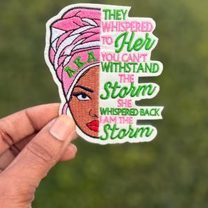 May include: An embroidered patch featuring a stylized illustration of a person wearing a pink headwrap with the letters AKA in green. The text reads: They whispered to her you can't withstand the storm she whispered back I am the storm.
