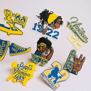 May include: A collection of embroidered patches in yellow, blue, and brown. The patches feature various designs, including sorority symbols, the number 1922, and illustrations of women. The patches are likely for apparel or accessories.