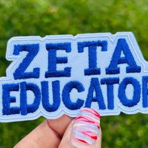 May include: A white embroidered patch with the words "ZETA EDUCATOR" in bold blue letters. The patch has a white border and is held against a blurred green background.
