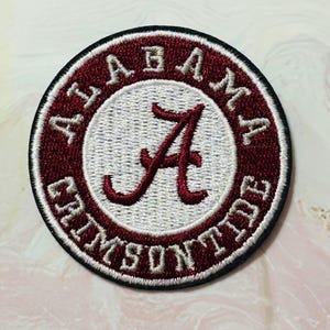 Puede incluir: Parche bordado circular con las palabras "ALABAMA CRIMSON TIDE" en letras blancas sobre un borde granate. El centro tiene un fondo blanco con una "A" granate.