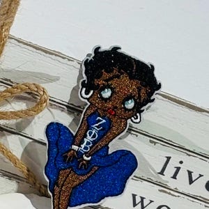 May include: A glittery Betty Boop-style pin. The pin features a cartoon woman in a blue dress with the word "Never" on it. The pin is set against a white background with the words "live worry be" visible.