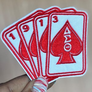 Puede incluir: Parches bordados con diseños de naipes. Los parches muestran los números 1, 9 y 3, junto con un símbolo de pica rojo y letras griegas. Los parches tienen un fondo blanco con detalles rojos.