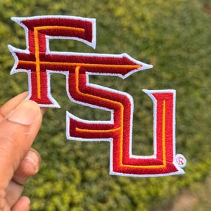 May include: An embroidered patch featuring the letters FSU in a stylized font. The letters are primarily red with a thin yellow inner border and a thick white outer border. A small registered trademark symbol is visible on the bottom right corner.