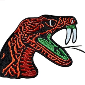 Puede incluir: Un parche bordado que muestra el perfil de una cabeza de serpiente con la boca abierta. El diseño utiliza hilo negro, naranja brillante, verde vibrante y blanco para representar las escamas, los colmillos y la lengua sobre un fondo negro.
