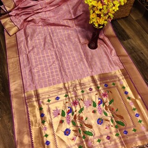 May include: A light pink silk saree with a gold border and intricate floral and peacock embroidery. The saree has a grid pattern and a decorative border with tassels. A vase of yellow flowers sits nearby.