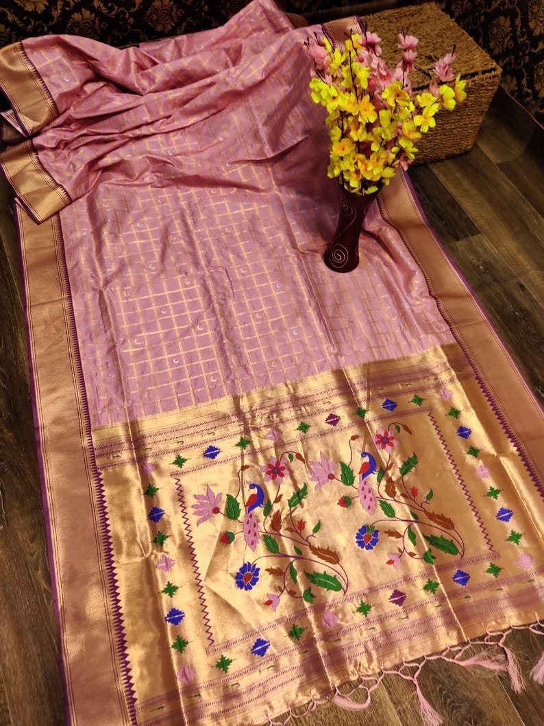 May include: A light pink silk saree with a gold border and intricate floral and peacock embroidery. The saree has a grid pattern and a decorative border with tassels. A vase of yellow flowers sits nearby.