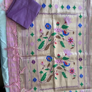 May include: A beige and pink textile with floral and geometric patterns. The fabric features a repeating design of flowers, birds, and geometric shapes in blue, green, and pink. A folded purple fabric square rests on top. The edges are trimmed with pink tassels.