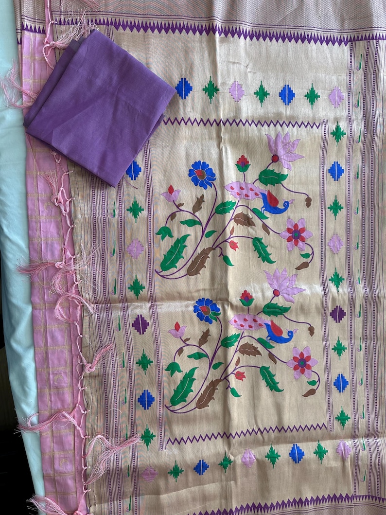 May include: A beige and pink textile with floral and geometric patterns. The fabric features a repeating design of flowers, birds, and geometric shapes in blue, green, and pink. A folded purple fabric square rests on top. The edges are trimmed with pink tassels.