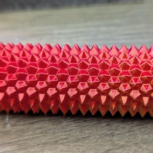 Puede incluir: Un objeto cil&iacute;ndrico rojo y naranja con una superficie texturizada y puntiaguda. El objeto tiene un patr&oacute;n geom&eacute;trico y un peque&ntilde;o extremo estriado de color naranja. El art&iacute;culo parece ser una herramienta de masaje o un objeto decorativo.