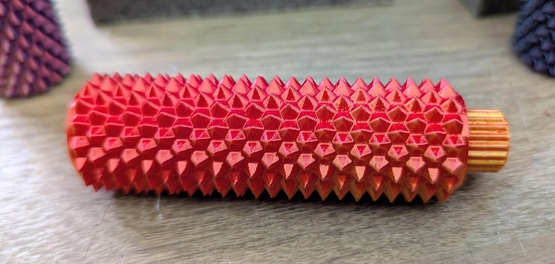 Puede incluir: Un objeto cil&iacute;ndrico rojo y naranja con una superficie texturizada y puntiaguda. El objeto tiene un patr&oacute;n geom&eacute;trico y un peque&ntilde;o extremo estriado de color naranja. El art&iacute;culo parece ser una herramienta de masaje o un objeto decorativo.