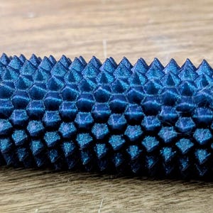 Puede incluir: Objeto cil&iacute;ndrico azul oscuro texturizado con una serie de p&uacute;as triangulares que cubren su superficie. El objeto tiene una peque&ntilde;a extensi&oacute;n estriada en un extremo. El art&iacute;culo est&aacute; sobre una superficie de madera.