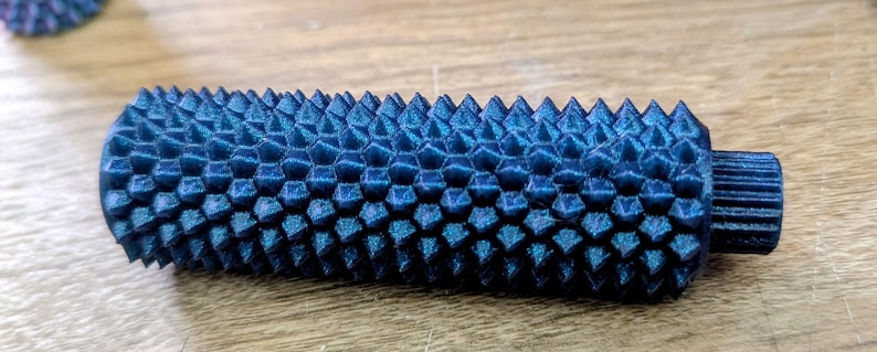 Puede incluir: Objeto cil&iacute;ndrico azul oscuro texturizado con una serie de p&uacute;as triangulares que cubren su superficie. El objeto tiene una peque&ntilde;a extensi&oacute;n estriada en un extremo. El art&iacute;culo est&aacute; sobre una superficie de madera.