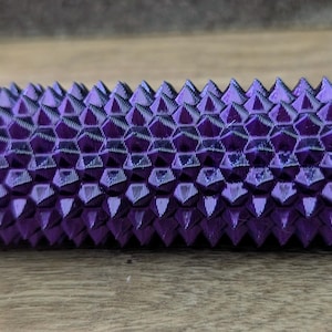Puede incluir: Un objeto cil&iacute;ndrico morado texturizado con un extremo azul. La superficie est&aacute; cubierta por un patr&oacute;n repetitivo de p&uacute;as triangulares. El objeto est&aacute; sobre una superficie de madera. Otros objetos similares son visibles en el fondo.