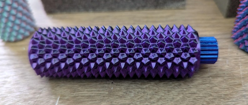 Puede incluir: Un objeto cil&iacute;ndrico morado texturizado con un extremo azul. La superficie est&aacute; cubierta por un patr&oacute;n repetitivo de p&uacute;as triangulares. El objeto est&aacute; sobre una superficie de madera. Otros objetos similares son visibles en el fondo.