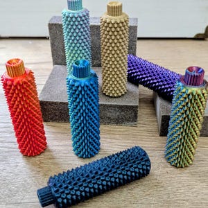 May include: A collection of colorful, textured cylindrical objects with a spiked surface. The objects are in various colors, including red, blue, green, and purple. The objects are arranged on a wooden surface and gray blocks.