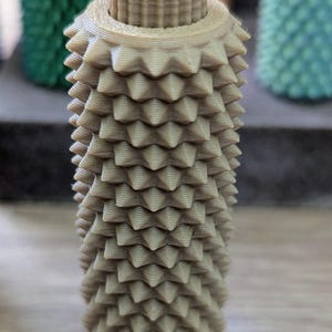 Puede incluir: Un tap&oacute;n de botella de color beige, impreso en 3D, con un dise&ntilde;o texturizado y puntiagudo. El tap&oacute;n tiene forma cil&iacute;ndrica con una parte superior estriada y una serie de protuberancias en forma de cono que cubren el cuerpo. Otros tapones coloridos son visibles en el fondo.