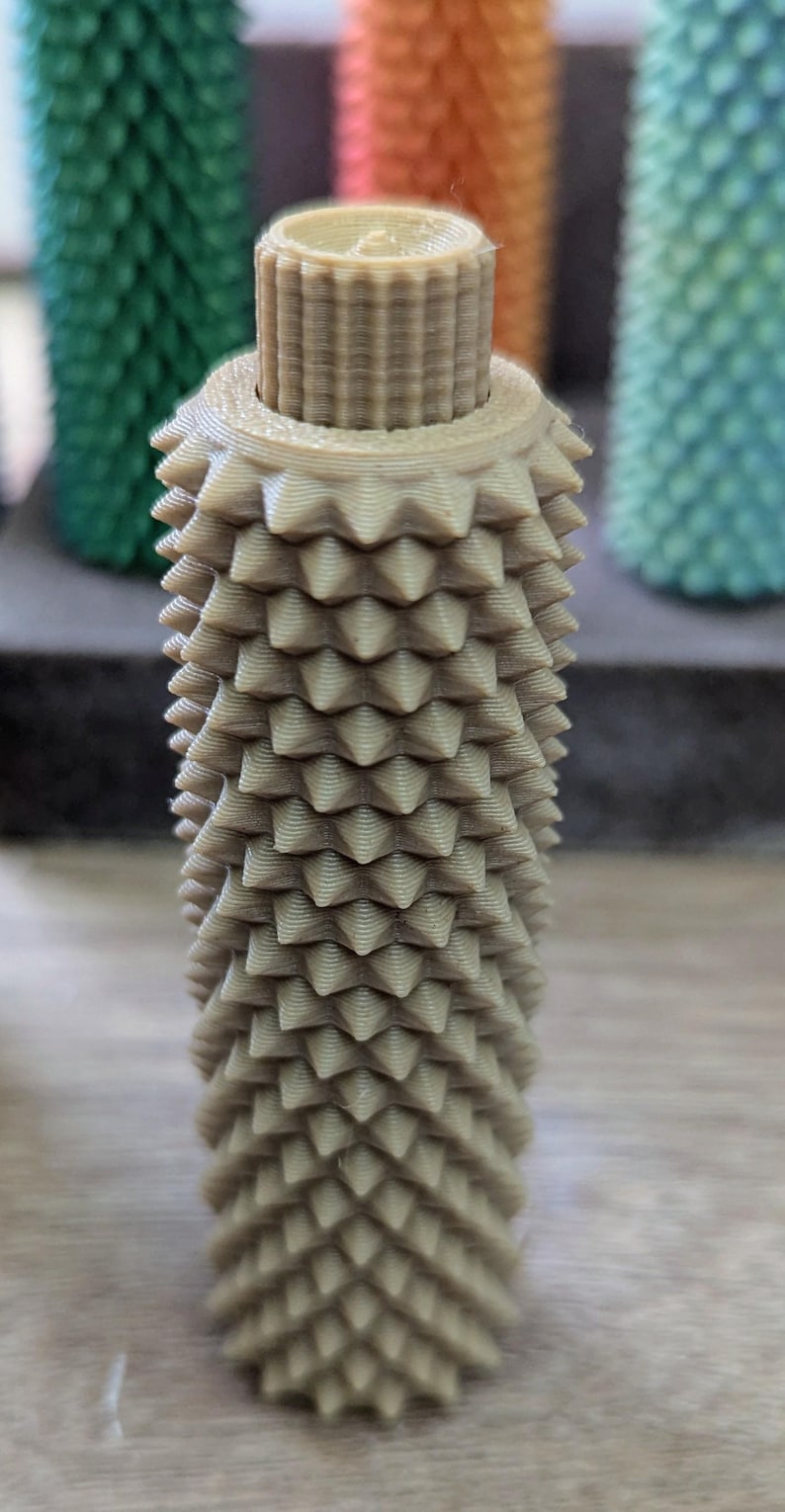 Puede incluir: Un tap&oacute;n de botella de color beige, impreso en 3D, con un dise&ntilde;o texturizado y puntiagudo. El tap&oacute;n tiene forma cil&iacute;ndrica con una parte superior estriada y una serie de protuberancias en forma de cono que cubren el cuerpo. Otros tapones coloridos son visibles en el fondo.