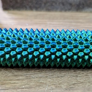 Puede incluir: Objeto cil&iacute;ndrico texturizado con una combinaci&oacute;n de colores verde azulado y azul. La superficie est&aacute; cubierta con un patr&oacute;n de p&uacute;as geom&eacute;tricas. El objeto tiene un extremo peque&ntilde;o y estriado. Otro objeto similar es visible en el fondo.