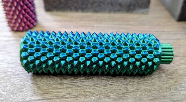 Puede incluir: Objeto cil&iacute;ndrico texturizado con una combinaci&oacute;n de colores verde azulado y azul. La superficie est&aacute; cubierta con un patr&oacute;n de p&uacute;as geom&eacute;tricas. El objeto tiene un extremo peque&ntilde;o y estriado. Otro objeto similar es visible en el fondo.